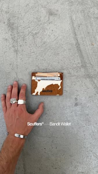Anuncio de Scuffers: mano con anillos junto a una cartera de piel sobre el suelo de cemento